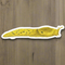 Watercolor banana slug sticker on wood surface, showcasing vibrant Pacific Northwest design. Durable vinyl, perfect for laptops or bottles. Created by artist Brigida Swanson, Yardia.