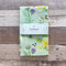Kitchen Garden Tea Towel - Organic Cotton Kitchen Towel, featuring a watercolor fruit and vegetable pattern, displayed on a wooden surface with a visible label.