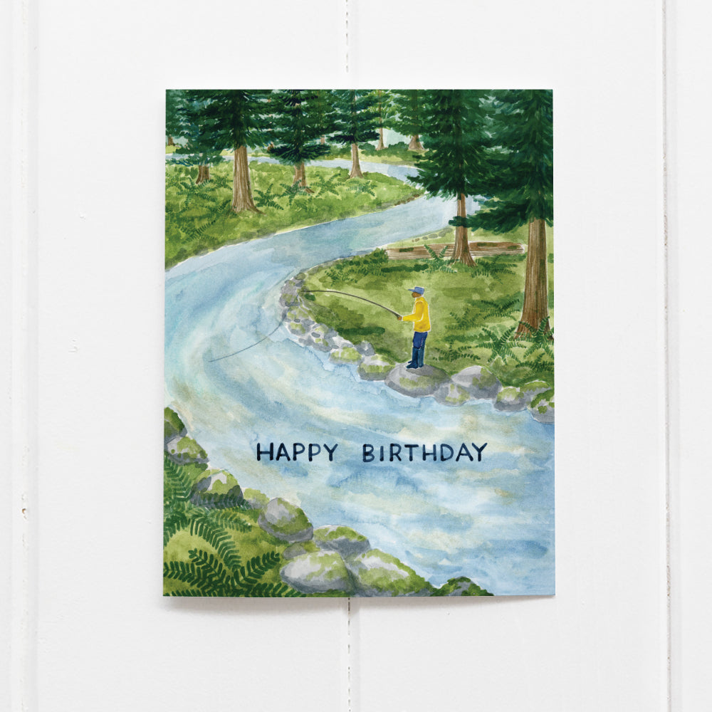 Fishing Birthday Card featuring a watercolor illustration of a fisherman in a trout river surrounded by forest, with a hand-lettered Happy Birthday message.