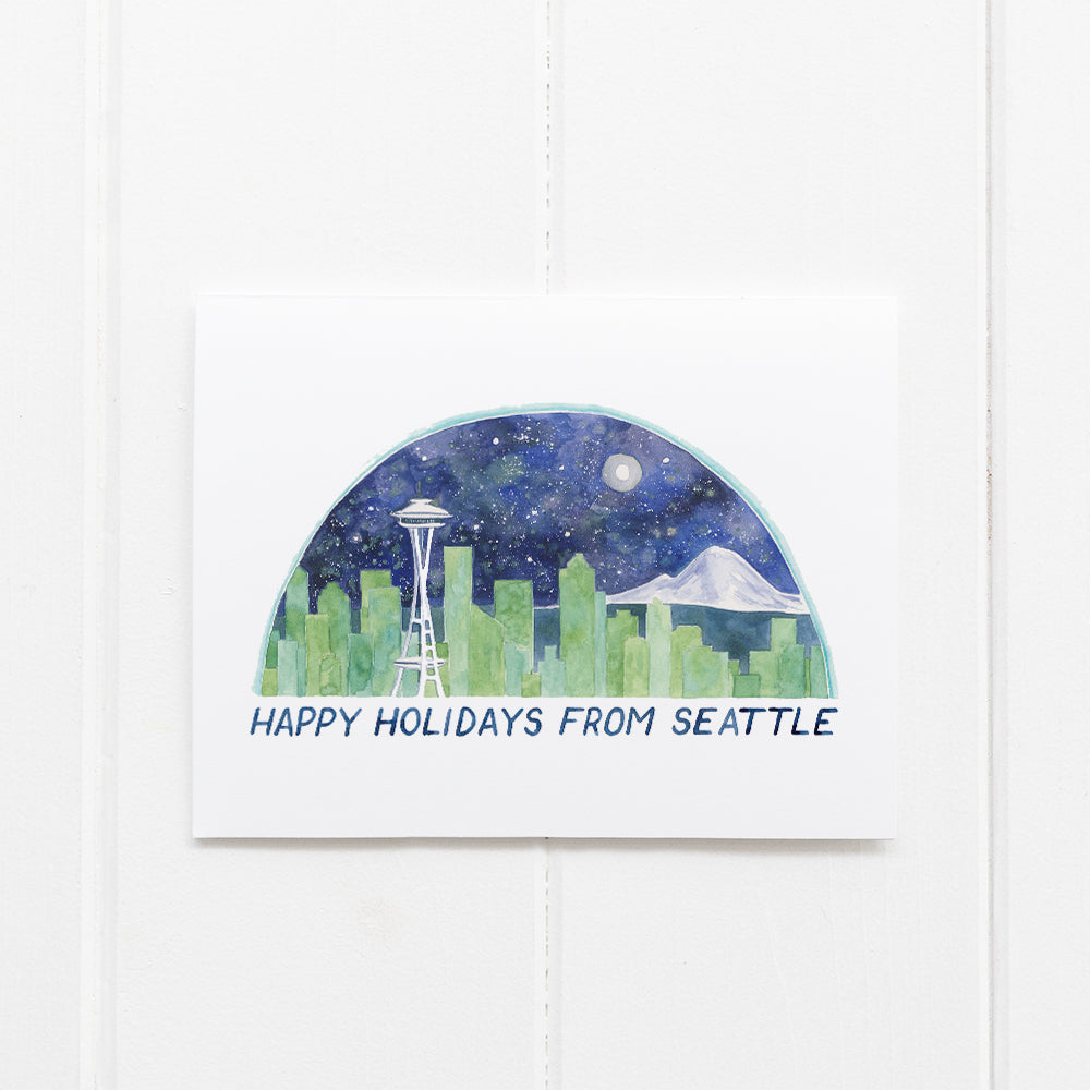 Seattle Boxed Holiday Cards - Set of 6, featuring a watercolor Seattle skyline with Space Needle beneath HAPPY HOLIDAYS FROM SEATTLE, propped against a white-paneled wall.