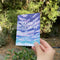 Hand holding a Season's Greetings Holiday Card featuring silver foil snowflake details and lettering, blank inside, with a kraft envelope.