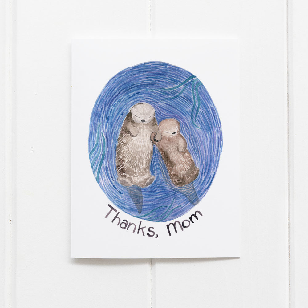 Thanks Mom Otters Card featuring a watercolor illustration of a mama and baby otter holding hands on recycled cardstock, blank inside, with a kraft envelope.