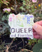 Hand holding a Queer And Flourishing Sticker, featuring watercolor wildflowers in rainbow order with hand-lettered text. Ideal for laptops or water bottles.