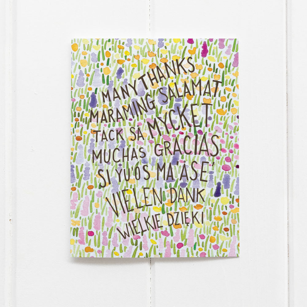 Multilingual Many Thanks Card featuring hand-lettered text in various languages, surrounded by a watercolor meadow illustration, designed by Brigida Swanson.