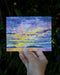 With Sympathy Card featuring a watercolor sunset off Seattle's Alki Beach, held in hand, showcasing Brigida Swanson's illustration. Blank inside, includes kraft envelope.