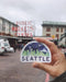 Seattle Sticker with a watercolor illustration of the Seattle skyline and Mt. Rainier, held in a hand outdoors, showcasing the city's iconic features.