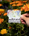 Hand holding the Multiracial and Flourishing vinyl sticker, featuring watercolor wildflowers and hand-lettered text, celebrating interracial love and multiracial identities.