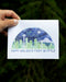 Seattle Boxed Holiday Cards - Set of 6, featuring a watercolor Seattle skyline with Space Needle and Happy Holidays from Seattle message, hand-held against out-of-focus foliage.
