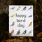 A square white greeting card with the words 'happy bird day' in dark brown lettering and illustrated birds around the border, placed on a bed of dry leaves and moss outdoors.