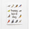 A white greeting card with the words 'happy bird day' in dark brown lowercase letters, surrounded by illustrated birds in multiple colors, photographed against a white wood plank background.