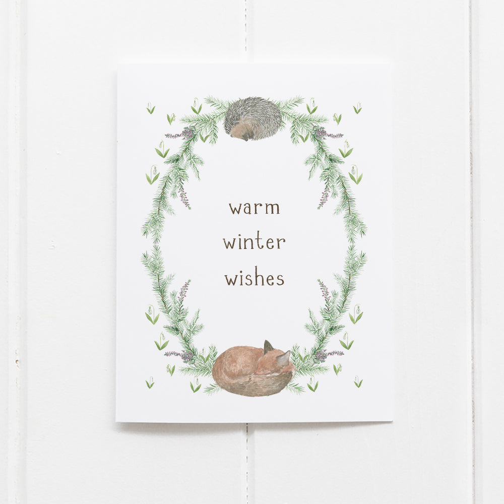 Winter Woodland Holiday Card displaying a watercolor wreath with a sleeping hedgehog and fox, surrounded by greenery, and warm winter wishes in hand-lettered text.