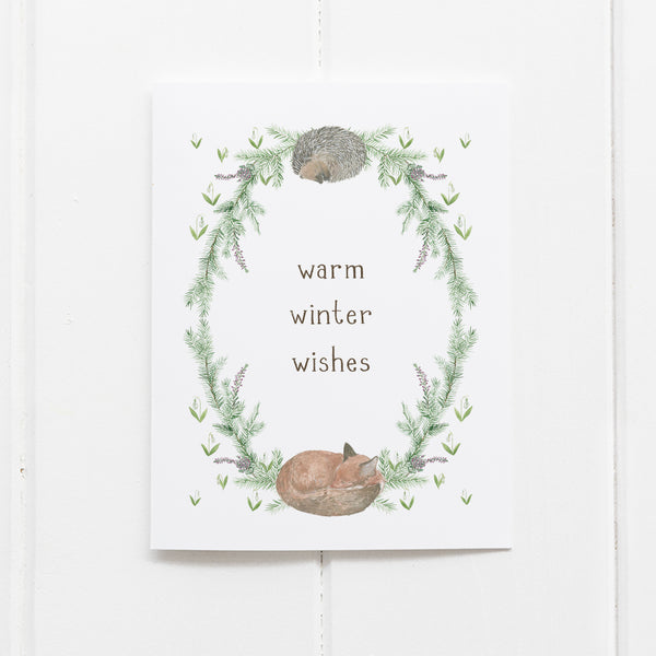 Winter Woodland Holiday Card displaying a watercolor wreath with a sleeping hedgehog and fox, surrounded by greenery, and warm winter wishes in hand-lettered text.