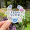 Trans Voter Sticker featuring watercolor floral design, held by a person, symbolizing LGBTQ+ pride and voter identity. Durable vinyl, weatherproof, 3x3.