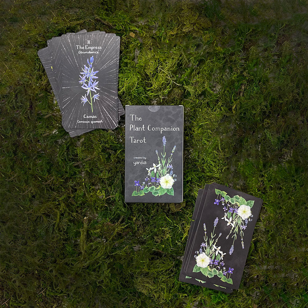 The Plant Companion Tarot Deck featuring watercolor botanical illustrations inspired by floriography, shown with floral-themed notebooks and a flower-adorned sign.