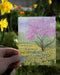 A hand holds a watercolor thank you card with illustration of cherry blossom tree and daffodils. The card reads sending you a garden of thanks.