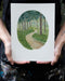 the artist holds her painting of a trail through a forest