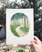 a hand holds a watercolor painting of a forested trail