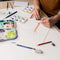 Image of the artist's hands painting a tree in watercolor on a messy desk