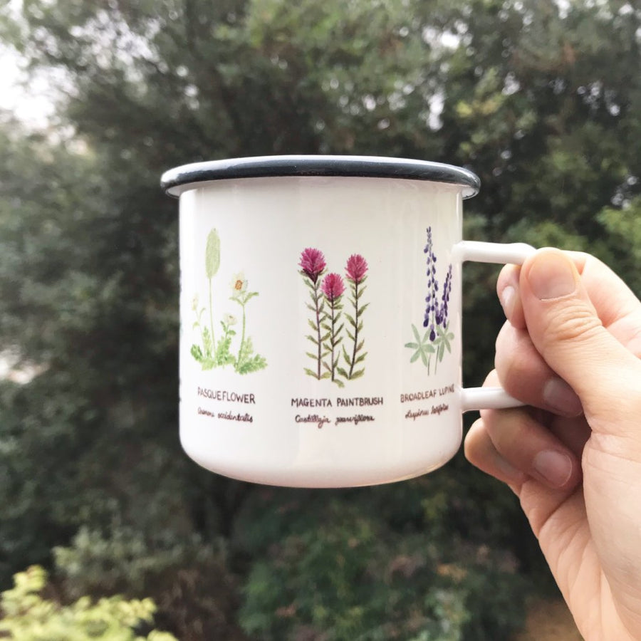 Hand holding a white enamel camp mug featuring Pacific Northwest wildflowers illustration, perfect for outdoor enthusiasts and cozy winter beverages.