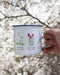 Hand holding a Wildflowers Camp Mug, a 12-ounce enamel mug with Pacific Northwest wildflower illustration, ideal for winter beverages or as a camping gift.